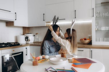 Sevgi dolu bir anne ve kız, anlamlı bir anı paylaşıyor el sanatları yaratıyor, Paskalya 'yı kutluyor ve parlak ve modern bir mutfakta kaynaşıyor. Tavşan kulağı ve renkli süslemeler gibi aksesuarlar şenlikli atmosferi vurguluyor.