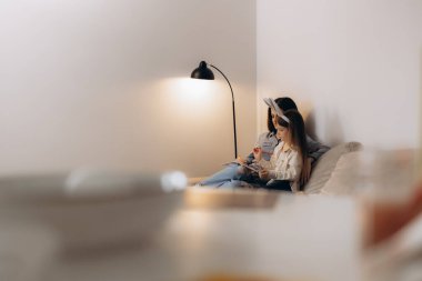 Bir annenin, minimalist bir ev ortamında, sıcak, parıldayan bir lambanın altında kızıyla kitap paylaştığı, birliktelik, rahatlık ve aile bağlarını çağrıştıran huzurlu bir an..