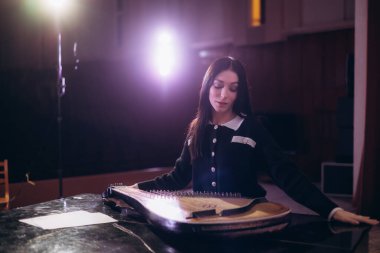 Odaklanmış stüdyo ışıkları altında geleneksel bir telli çalgıyla müzik yapan genç bir kadın yaratıcılığın huzurlu ve sanatsal atmosferini yakalıyor..