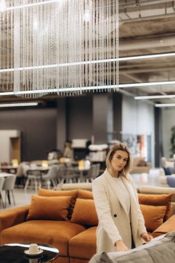 Bej ceketli zarif bir kadın modern iç mekanlarla çevrili bir mağazada rahat mobilyaları keşfediyor..