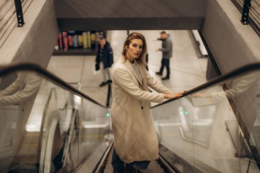 Bej ceket giyen şık bir kadın şık bir şehir ortamında yürüyen merdivenden iner. Sahne, kentsellik, moda ve kamu altyapısına vurgu yapan modern yaşam tarzı kavramlarını yansıtıyor..