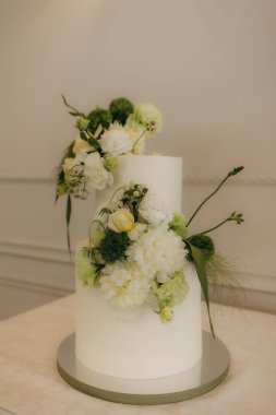 Yumuşak yeşil ve beyaz tonlarda çiçeklerle süslenmiş güzel tasarlanmış beyaz bir düğün pastası özel bir kutlama için mükemmel, zarif ve romantik bir ortam yaratıyor..