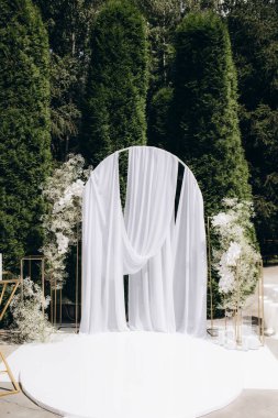 Akan beyaz kumaş, gür yeşillik ve narin çiçek düzenlemeleri ile süslenmiş güzel bir açık hava düğün kemeri, sakin bir ortamda uzun yeşil ağaçlarla çevrili bir zarafet ve romantizm atmosferi yaratıyor..