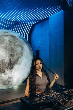 Bir DJ, parlak ışıklarla aydınlatılmış yaratıcı bir alanda, bir konsolda müziği Ay benzeri bir kürenin arka planına karşı karıştırarak büyüleyici ve ilham verici bir atmosfer yaratır..