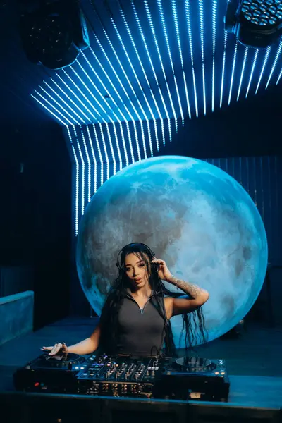 Bir kadın DJ, büyük bir ay arkaplanı ve neon ışıklandırma tasarımlarının yer aldığı fütürist ve canlı bir ortamda performans sergiliyor. DJ stili bir mikser işletiyor, kendine güven ve tarz yayıyor..