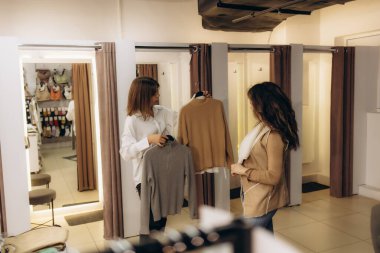 Rahat bir butik mağazasında, modern prova odaları ve şık giysi parçalarının bulunduğu süveter seçeneklerini araştıran iki kadın..