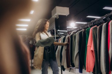 Bir kadın modaya uygun bir mağazanın rafındaki çeşitli kıyafetlere zevkle göz atıyor. Atmosfer, keyif, moda ve tüketiciliği yansıtıyor..