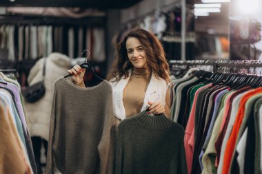 Neşeli bir kadın rahat bir giyim mağazasında alışveriş yaparken süveterleri inceliyor, moda tercihlerini gösteriyor..