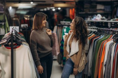 İki arkadaş, şık giysilerle çevrili modern bir giyim mağazasında alışveriş yaparken mutlu bir şekilde sohbet ediyorlar. İfadeleri, paylaşılan deneyimlerin ve moda keşiflerinin neşesini yansıtıyor..