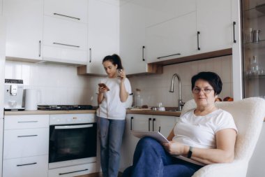 İki kadın mutfakta vakit geçiriyor, biri oturuyor ve yazıyor, diğeri de akıllı telefon kullanıyor, modern yaşam tarzlarını ve etkileşimlerini gösteriyor..
