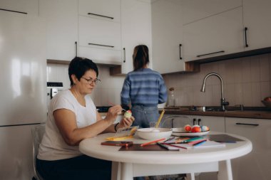 A mother and daughter engaged in egg decorating. Modern kitchen decor offers bright and lively ambiance. The activity encapsulates bonding and creativity in a family setting. clipart