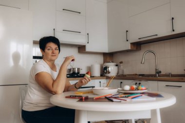 Woman painting and decorating Easter eggs at a table in a clean modern kitchen, accompanied by art supplies such as sketches and colored pencils, showcasing a moment of creative seasonal preparation. clipart