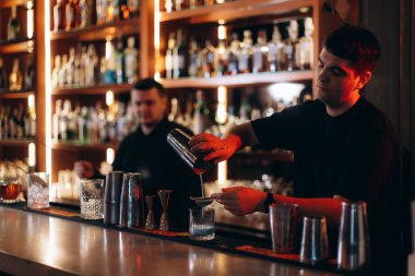 Yetenekli bir barmen, bir meslektaşının yanında bardağa kokteyl doldurur. İçkilerle dolu şık bir bara karşı hazırlanır. Bu resim canlı ve modern bir bar atmosferini gösteriyor..