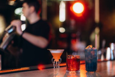 Tezgahında renkli kokteyller olan canlı bir bar atmosferi. Bir barmen içkileri özenle aydınlatır, sofistike ve sosyalleşmeyi çağrıştırır..