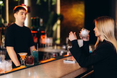 Modern bir barda genç bir bayanın içkisinin keyfini çıkardığı dostane bir atmosfer. Sıcak bir aydınlatma ve şık bir dekorun ortasında barmenle etkileşim halinde..