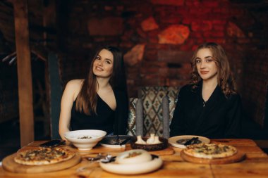 Ahşap bir masada oturan iki arkadaş, sıcacık, kırsal bir restoranda yemek yiyorlar. Ortam aydınlatma ve zarif dekor anlamlı konuşmalar ve lezzetli yemek deneyimleri için mükemmel sıcak bir atmosfer yaratıyor..