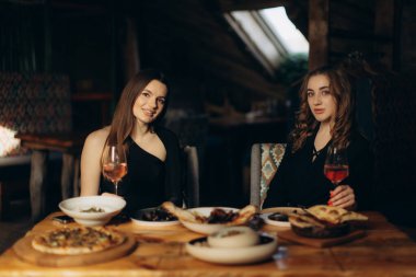 İki arkadaş, sıcak ve sıcak bir restoranda güzel bir yemek yiyorlar. Her biri, ahşap bir masada sunulan lezzetli yiyeceklerle çevrili bir içecekle bardakları tutuyor..