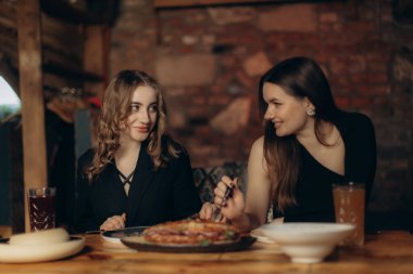 İki dişi bir yemeği paylaşıyor ve sıcak bir şekilde gülümsüyor. Büyüleyici bir restoran ortamında, ahşap bir masada oturuyorlar. İfadeleri ve çevreleri sıcaklığı, dostluğu ve arkadaşlığın neşesini iletir..