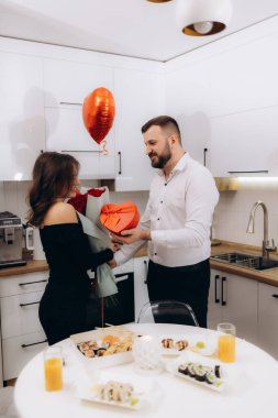 Güzel bir mutfakta romantik bir yemeğin tadını çıkaran bir çift, hediye alışverişi yapıyor ve kutlamanın sıcaklığıyla çevrili hassas anları paylaşıyor..