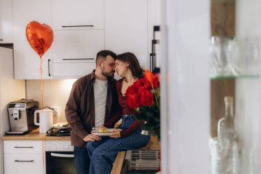 Sevgi dolu bir çift, kalp şeklinde bir balon, bir buket kırmızı gül, şefkat ve sıcaklık gibi romantik unsurlarla çevrili, samimi bir mutfakta kucaklaşır..
