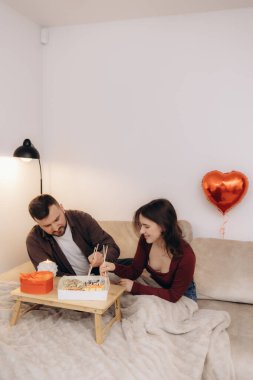 Sevgi dolu bir çift romantik unsurlarla süslenmiş rahat bir yatakta suşiyi paylaşır. Kırmızı kalp şeklinde bir balon, dingin ortama sıcak ve samimi bir cazibe katar..