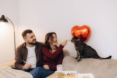 Kanepede oturan mutlu bir çift, arka planda kırmızı kalp şeklinde bir balonla güçlendirilmiş sıcak, samimi bir ev ortamında meraklı kedileriyle suşi paylaşıyorlar..
