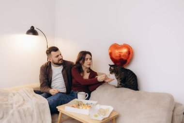 Bir çift sıcak kanepelerinde oturup sushi yiyor, yanında sevimli kedileri ve kalp şeklinde bir balon ile birlikte sevgi, birliktelik ve rahatlama hissi yayıyorlar..