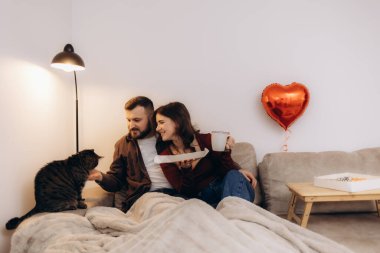 Bir çift evde kedileriyle rahat bir anın tadını çıkarıyor, kahve ve atıştırmalıkları paylaşıyor, sembolik bir kalp şeklindeki balonla süsleniyor, sıcak ve sevgi dolu bir atmosfer yaratıyor..