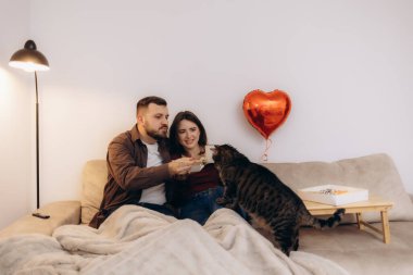 Bir çift, neşeli kedileri ve dekoratif kırmızı kalp balonlarıyla birlikte kanepede rahat bir anın tadını çıkarıyor. Sıcak ve sevecen bir atmosfer yaratıyorlar..