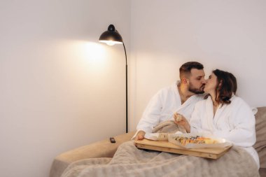 Bornozlu sevgi dolu bir çift sıcak lamba ışığı altında sıcak bir evde birlikte sushi içerken aynı hassas anı paylaşıyor..