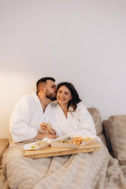 Mutlu bir çift bornozla birlikte rahatlar, rahat bir kanepede suşi yer, romantik ve rahatlatıcı bir akşamın tadını çıkarır. Huzurlu evlerinde sevgi, rahatlık ve beraberlik sergilerler..