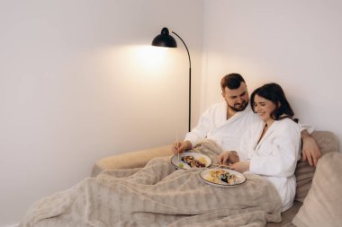 Beyaz bornoz giymiş sevgi dolu bir çift sıcak bir lambanın altındaki kanepede suşinin tadını çıkarıyor..