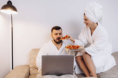 Bornozla rahatlayan, pizzalarını paylaşan ve rahat evlerinde dizüstü bilgisayar kullanan bir çift. Rastgele ve eğlenceli bir an. Rahatlık, sevgi ve rahatlık..