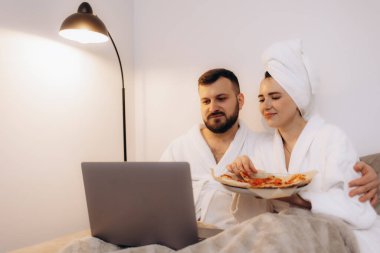 Bornoz giyen bir çift pizzayı ve dizüstü bilgisayarda film izleyerek rahat ve rahat bir atmosfer yaratıyor..