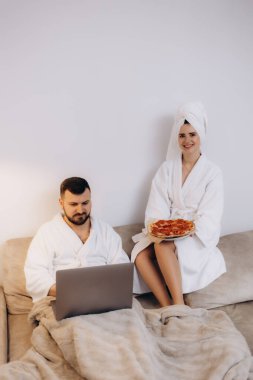 Bornoz giyen bir çift kanepede rahatlar, biri dizüstü bilgisayar kullanır, diğeri pizza tutar, evde rahat ve rahat bir anın tadını çıkarırlar..