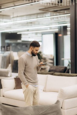 Genç adam parlak, modern bir mobilya dükkanında akıllı telefonuna bakıyor. Sahne, şık bir alışveriş ortamında kişinin rahat ve düşünceli anını yakalar..