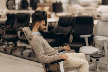 Modern koltuklarla çevrili bir mobilya mağazasında ergonomik ofis sandalyesini test eden bir adam. İş yeri mobilyaları, konfor ve ofis malzemeleri için mükemmel bir görsel.