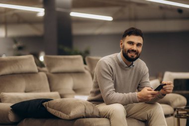 Akıllı telefon tutan sıradan giyinmiş bir adam, rahat bir kanepede oturuyor rahat ve neşeli bir tavırla, modern bir ev ortamında..