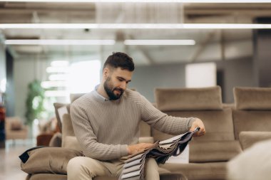 Sakallı adam şık bir dükkanda zarif bir koltukta oturur ve kumaş örneklerini dikkatlice inceler..