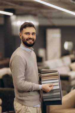 Modern bir mağazada elinde kumaş örnekleri olan, tasarım ve seçim çeşitlerini sergileyen bir adam. Sahne, dost canlısı müşteri hizmetlerini ve iç tekstil seçimini iyi aydınlatılmış bir ortamda yansıtıyor..