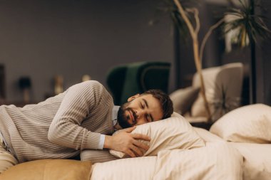 Süveter giyen sakallı bir adam bir yatakta rahat bir şekilde dinleniyor, rahatlık ve sükunet yayıyor..