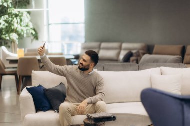 Şık bir kanepede oturan bir adam parlak ve modern bir oturma odasında selfie çekiyor..