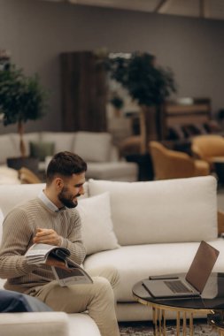 Modern beyaz koltukta oturan bir adam şık bir oturma odasında, elinde bir dergi tutarken dizüstü bilgisayarla çalışıyor, birden fazla iş yapma, üretkenlik ve çağdaş yaşam tarzı senaryosu öneriyor..