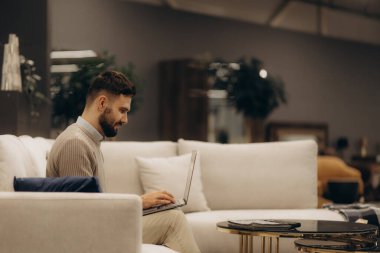 Sakallı bir adam, modern ve rahat bir oturma odasında, rahat bir atmosferi ve şık dekorasyonu olan bir dizüstü bilgisayar kullanarak beyaz bir kanepeye rahatça oturur..
