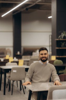 Çağdaş bir mobilya galerisinde, çeşitli modern ve şık sandalye tasarımları sergileyen, samimi bir adam gülümsüyor. Müşteri etkileşimiyle mobilyacı ortamının mükemmel tasviri.