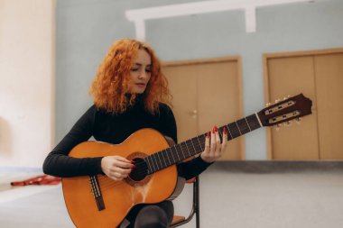 Kıvırcık kızıl saçlı, evde akustik gitar çalan, müziği ve sanatsal ruhu ön plana çıkaran genç bir kadın. Sahne, dingin ve modern bir ortamda yaratıcılık ve adanmışlığı yakalıyor..