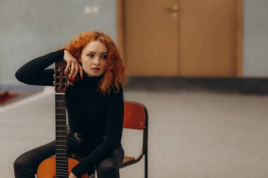 Sessiz bir stüdyoda akustik gitarla oturan kızıl saçlı kadın yaratıcılık ve sanatsal ifadeyi canlandırıyor. Siyahlar içinde, bir iç mekan ortamında özgüven ve tefekkür sergiliyor..