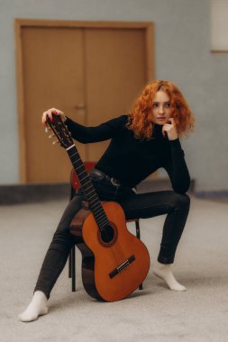Kıvırcık saçlı, sıradan giyinmiş genç bir kadın, kapalı bir yerde oturmuş klasik bir gitarla kendine güvenerek poz veriyor, bireyselliğini ve sanatsal ifadesini minimalist bir ortamda sergiliyor..