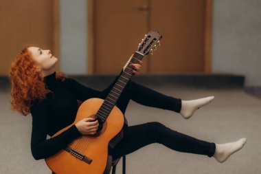 Siyah giyinmiş kızıl saçlı bir kadın, sanatsal duruşu, yaratıcılığı, bireyselliği ve müzikal tutkusunu yansıtan kapalı mekanda akustik gitar çalıyor..