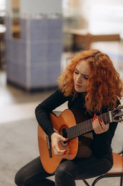 Kızıl kıvırcık saçlı bir kadın rahat bir ortamda odaklanmış ve ifade edilmiş bir gitar çalar. Sahne, samimi bir ortamda müzik tutkusu, yaratıcılık ve kişisel ifade temaları çağrıştırıyor..